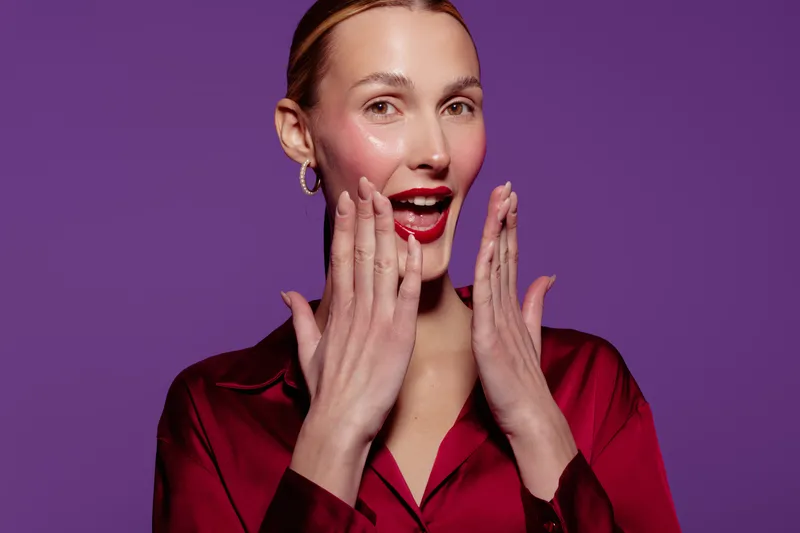 Surprised woman with red lipstick and earrings touches her face with both hands on purple background. Concept of beauty, skincare, makeup, and female emotions in studio lighting.