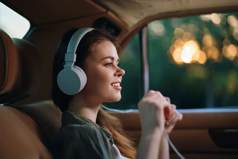 Young woman enjoying music with headphones in a car, smiling and relaxed, capturing the essence of joy and freedom during a sunny day.