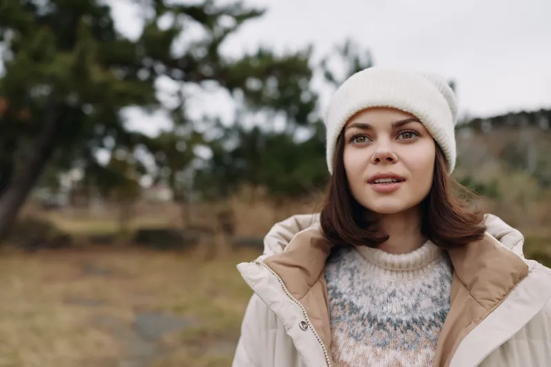 Woman in outdoor setting wearing a beanie, cozy sweater and light jacket, exploring a chilly landscape with distant trees.