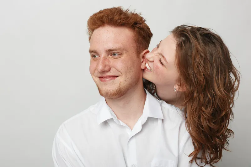 Happy young couple with red hair and curly brown hair share a joyful moment, smiling and embracing against a plain light background. Concept of love, friendship, and happiness.