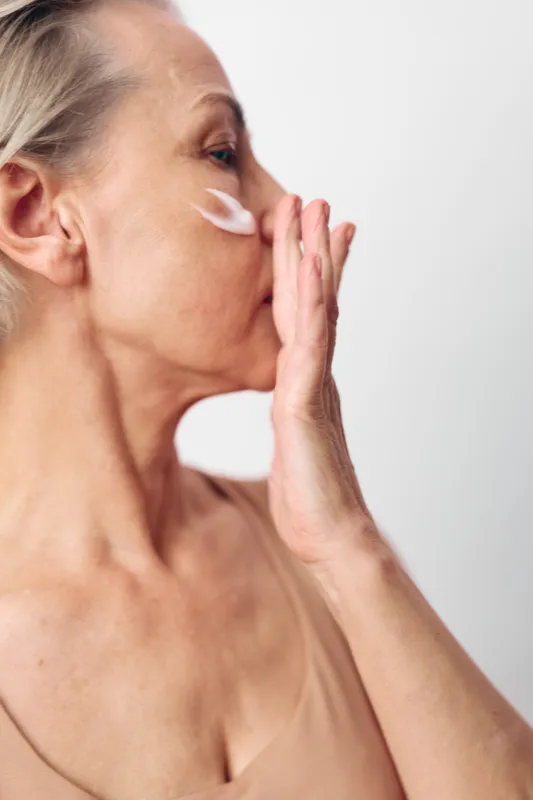 Mature woman applying cream on her cheek with hand near mouth, close-up on white background. Concept for skincare, anti aging, facial care, and natural beauty routine.