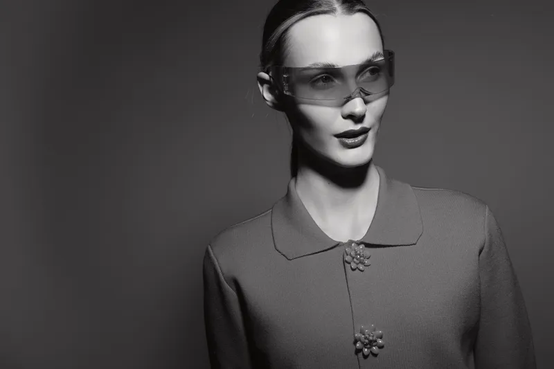 A young Caucasian woman with fair skin and slicked-back hair wears futuristic clear glasses and a textured jacket, posing against a dark background. Concept of fashion and modern style.