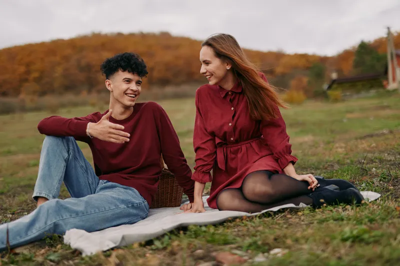 [A genuine candid moment of two friends sharing a warm, authentic conversation during an autumn picnic, capturing natural smiles, relaxed posture, and believable emotional connection.]