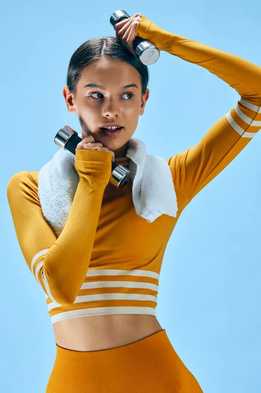 Female athlete in bright orange sportswear lifts dumbbells with a towel around her neck, against a vivid blue backdrop, showcasing strength, focus, and dynamic fitness of training.