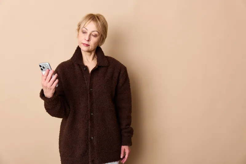 Elderly woman looks worried holding a smartphone, fingers near the screen. Studio portrait on a beige background, concept of technology stress and privacy.