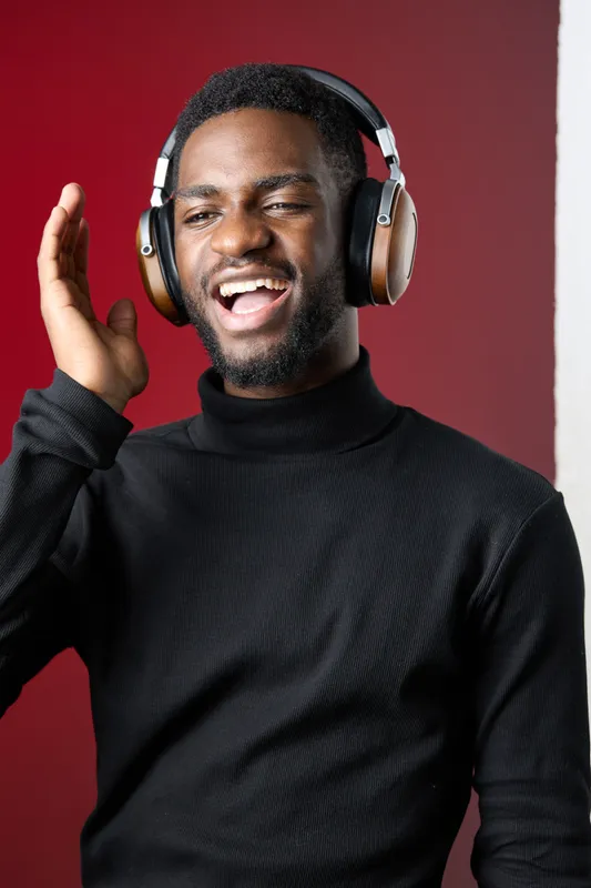 A joyful young Black man in a black turtleneck wearing headphones, smiling and enjoying music against a vibrant red background