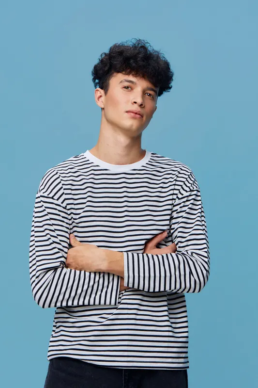 young man wearing striped long sleeve shirt posing with arms crossed against solid blue background, casual style and confident expression, trendy haircut
