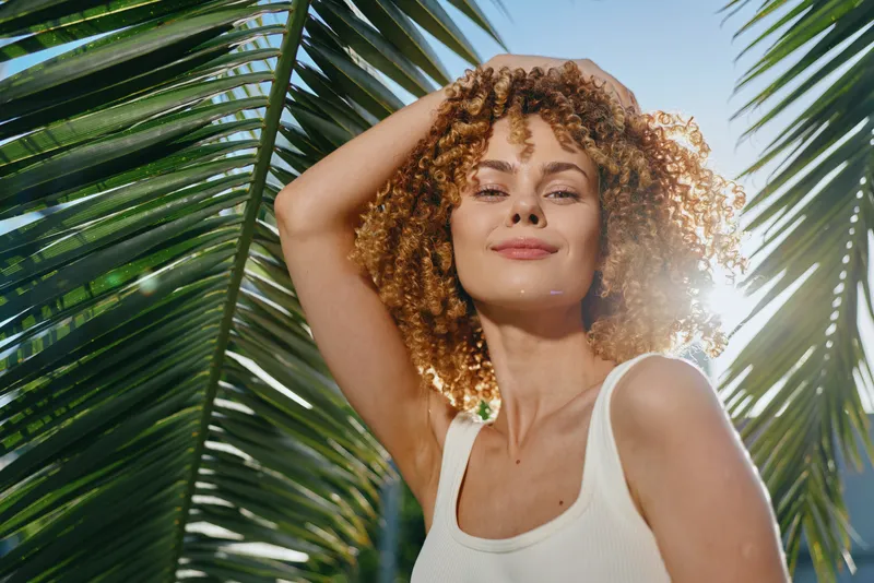 Smiling woman with curly hair enjoys tropical sunlight under palm leaves, expressing happiness and warmth in a bright summer setting.