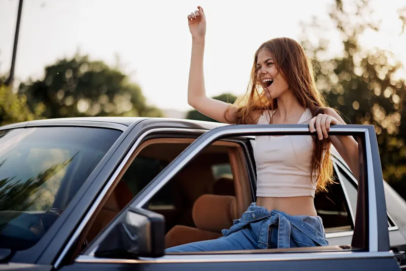 Young woman joyfully leaning out of car window, celebrating freedom and adventure in summer sun, wearing casual clothing with a vibrant background.