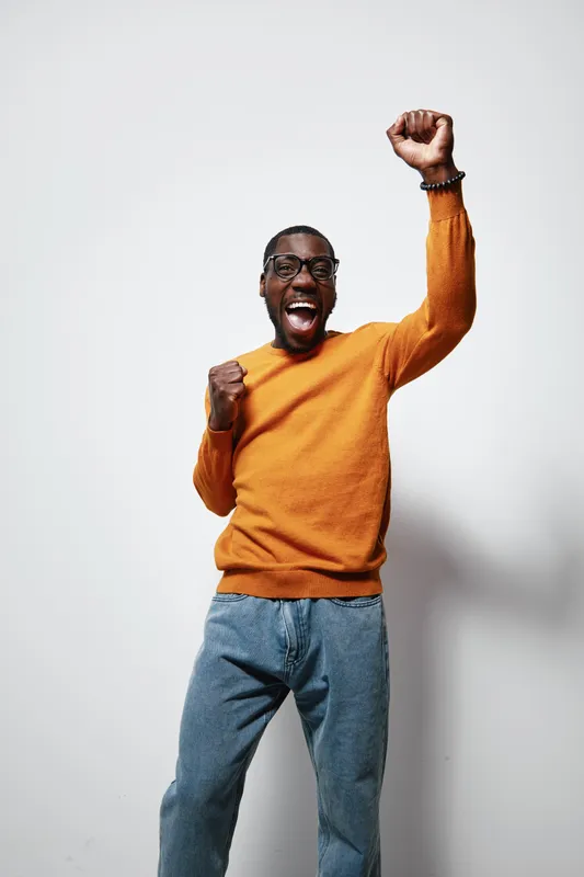 African American man with dark skin wearing orange sweater and blue jeans celebrating with raised fist and joyful expression on white background. Casual style and happiness.
