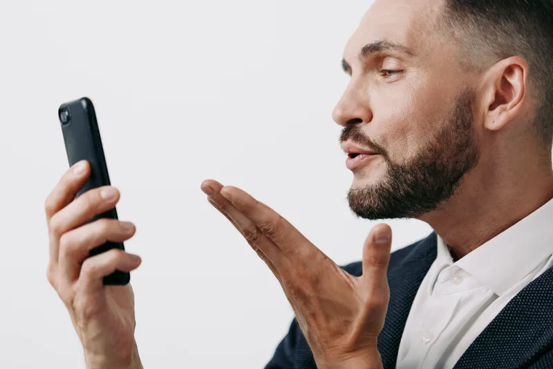 A professional man in a suit speaks into a smartphone, showing focus and confidence against a clean colored background, an isolated setting that highlights his business persona and emotion.