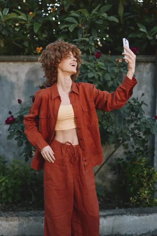 Curly haired woman in a rust suit takes a casual selfie outdoors, wearing a yellow crop top, with leafy greenery behind and warm natural light creating a relaxed mood
