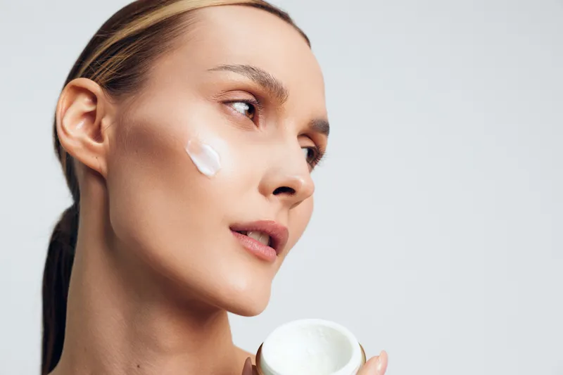 A young woman applies white cream on her cheek while holding an open jar, demonstrating skincare routine and facial care in a clean, bright environment.