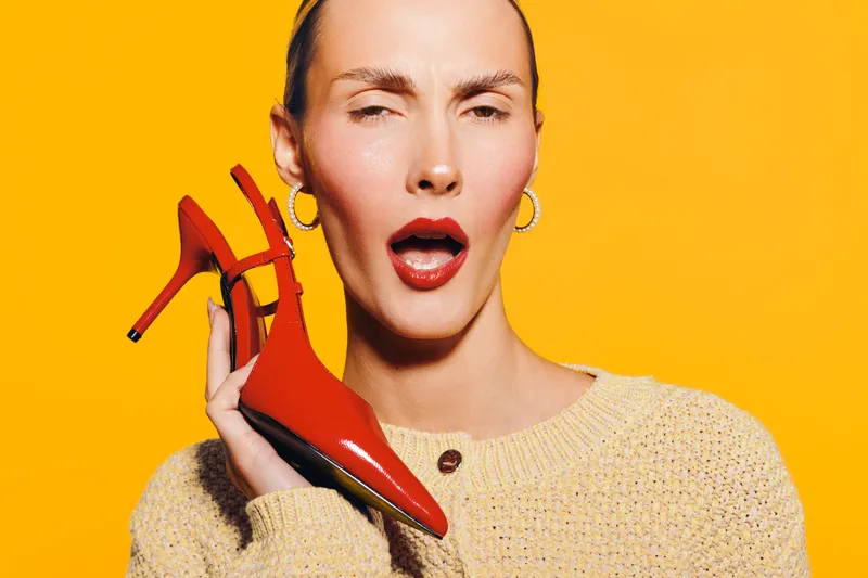 Young woman with red high heel shoe near face, wearing beige sweater and hoop earrings, posing on bright yellow background. Concept of fashion, style, and bold accessories.