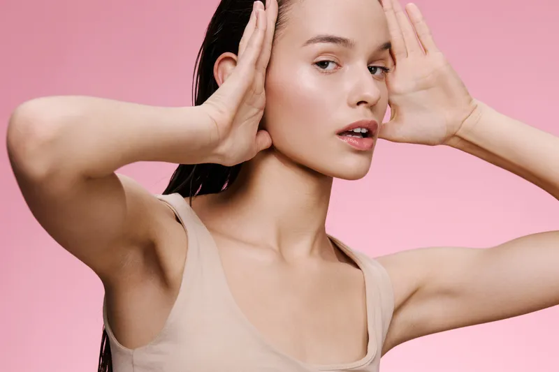 Young woman with long hair in a nude tank top, expressing calmness against a soft pink background, emphasizing beauty and self care concepts