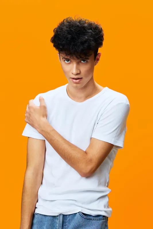 young man with curly hair wearing white t-shirt stands against bright orange background, showing casual style and natural expression in vibrant studio setting