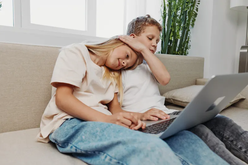 Two children sitting on sofa using laptop together, girl leaning on boy, casual home interior with natural light and plant behind.