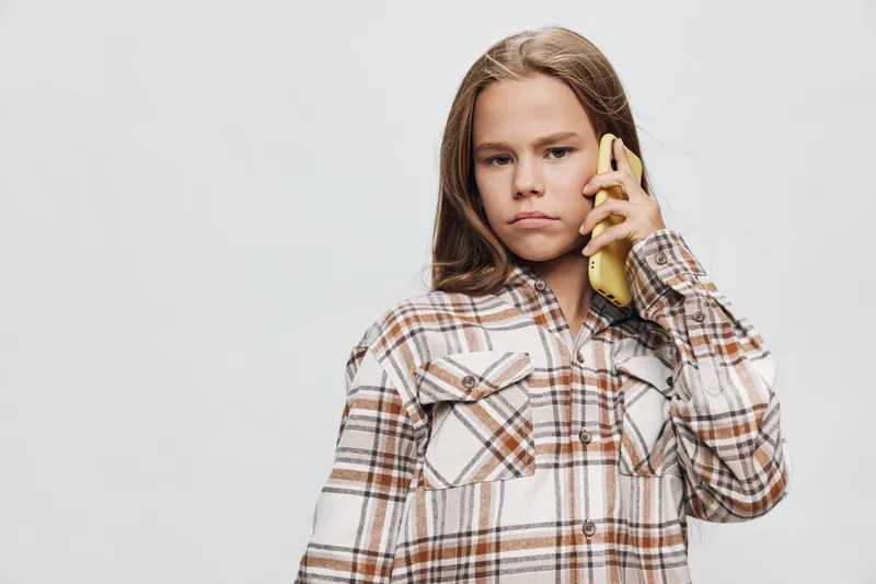 Teenage girl with long hair wearing checkered shirt talking on phone with serious expression on plain background studio shot. Communication, youth and technology concept.