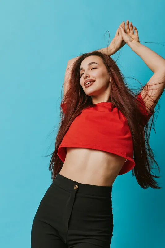 Young woman with long hair wearing red crop top and black pants posing with arms raised against blue background. Casual fashion, confident smile, and relaxed mood in studio portrait.