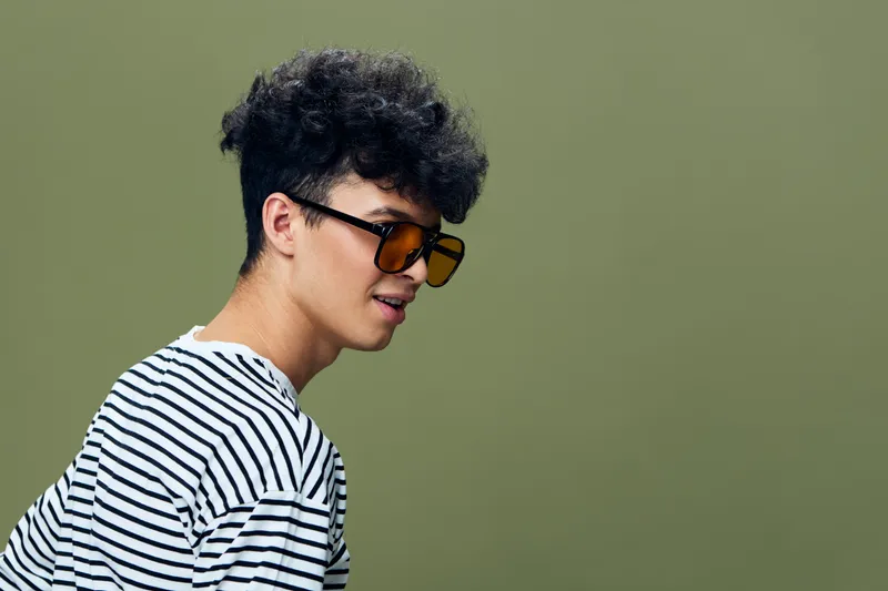 young man with curly hair wearing striped shirt and sunglasses, casual style, olive green background, fashionable look, studio portrait photo