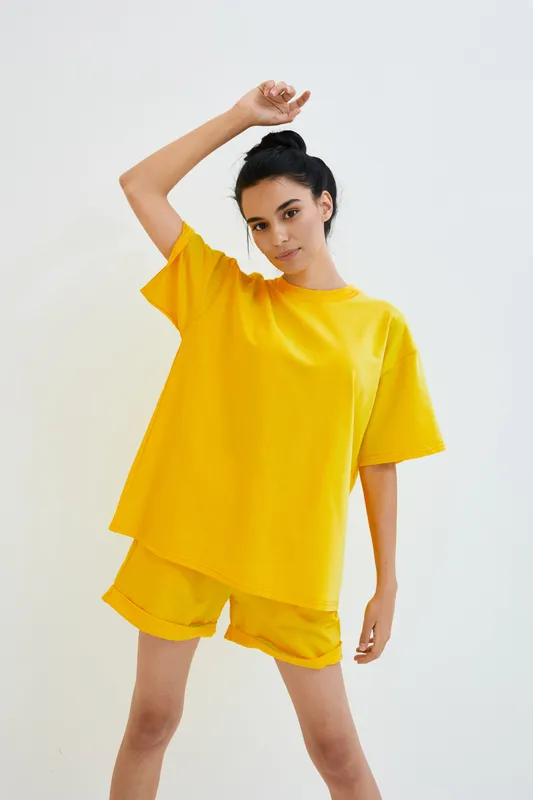 Young woman in bright yellow casual t-shirt and shorts stands confidently against plain white background, exuding a relaxed and modern style with her natural look.