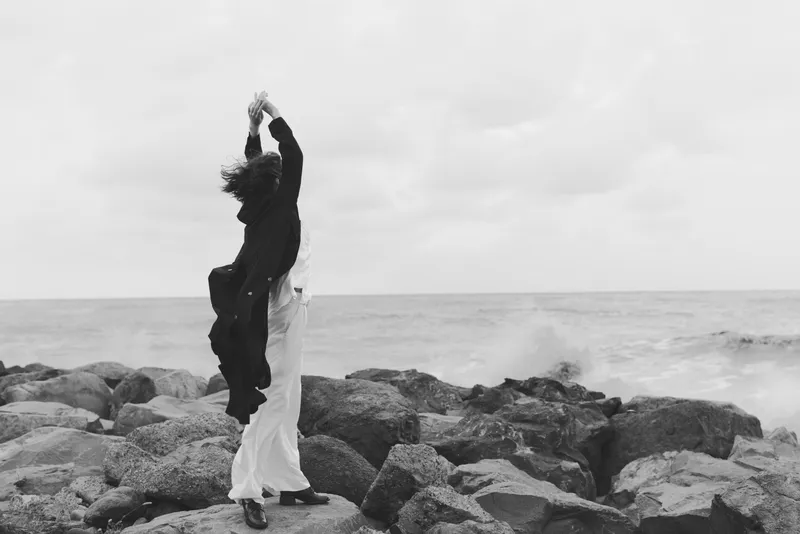 Couple dancing on a rocky shoreline by the sea, arms raised, flowing garments in motion, monochrome coastal moment evoking romance, celebration, and freedom against crashing waves and open horizon