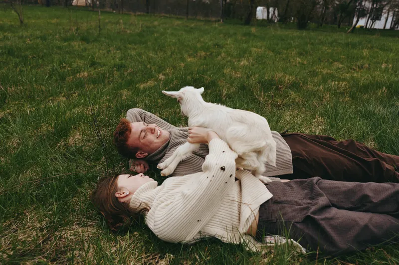 Lifestyle moment with a couple lying on grass outdoors, a goat kid nearby, they smile and embrace, rustic calm scene, soft lighting, candid everyday joy, harmony between humans and animals.