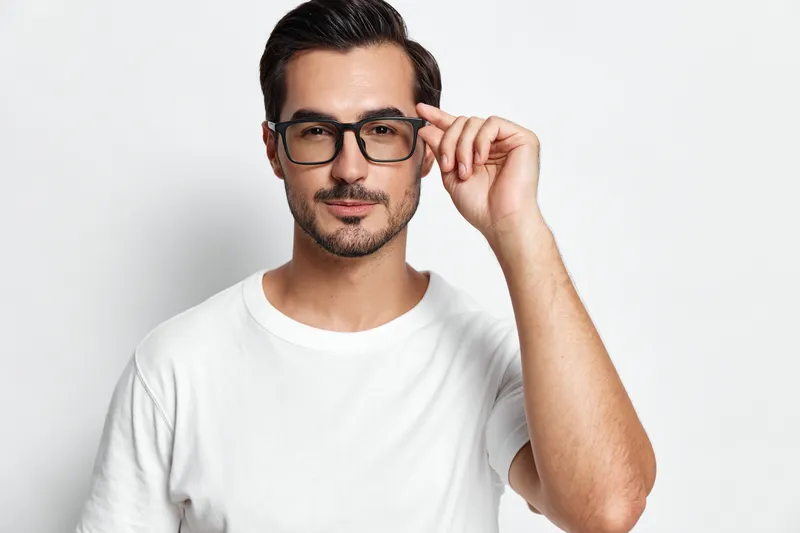 Stylish young man adjusting his glasses, looking directly at the camera with a confident expression. He is wearing a casual white t-shirt on a plain background.