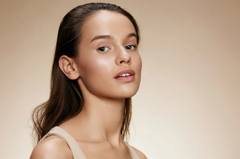 Natural beauty portrait of a young model with light brown hair and soft skin, showcasing a fresh makeup look against a beige background, perfect for beauty marketing