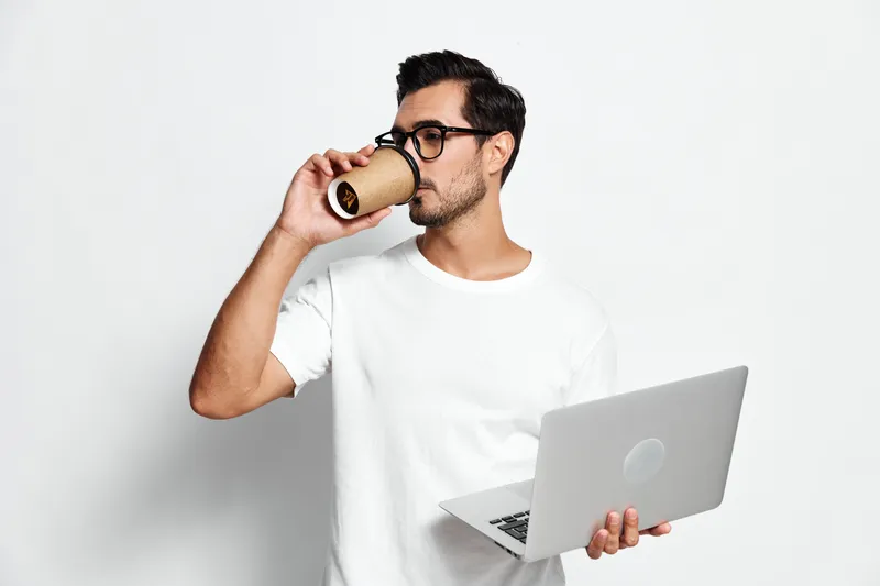 Young handsome man wearing glasses holding a laptop in one hand and a disposable coffee cup in the other, looking thoughtful and engaged against a light background.