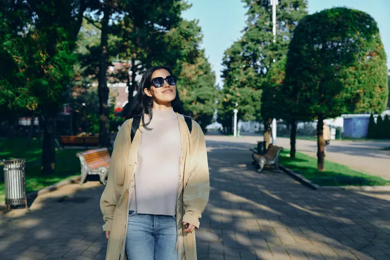 Woman walking in a sunlit park wearing a beige coat, jeans and sunglasses, relaxed pose amid trees and benches on a paved path, capturing casual outdoor style and fresh air