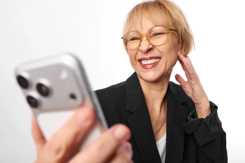 Woman smiling with phone indoors, wearing glasses and business blazer, communication call gesture, close up portrait, bright white background, soft light, relaxed mood