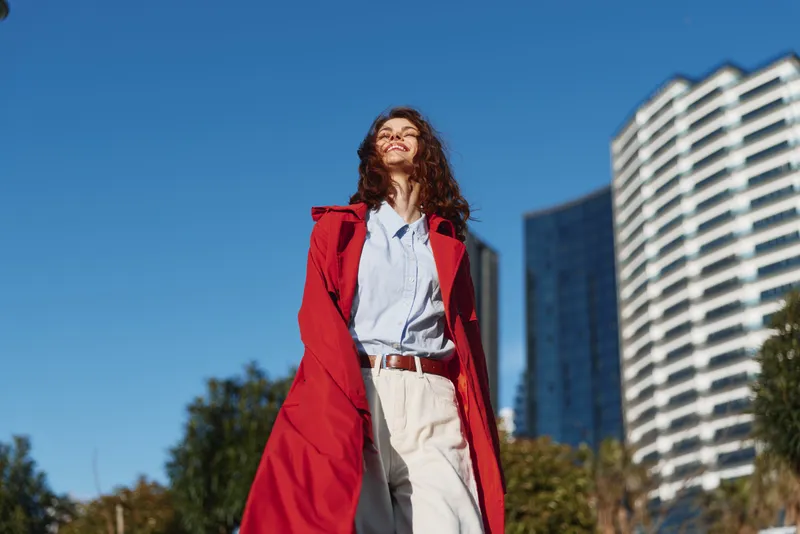 Bright urban portrait of a woman wearing a red coat, light trousers, and blue shirt, standing outdoors with skyscrapers, sunshine and trees, conveying confident street fashion.