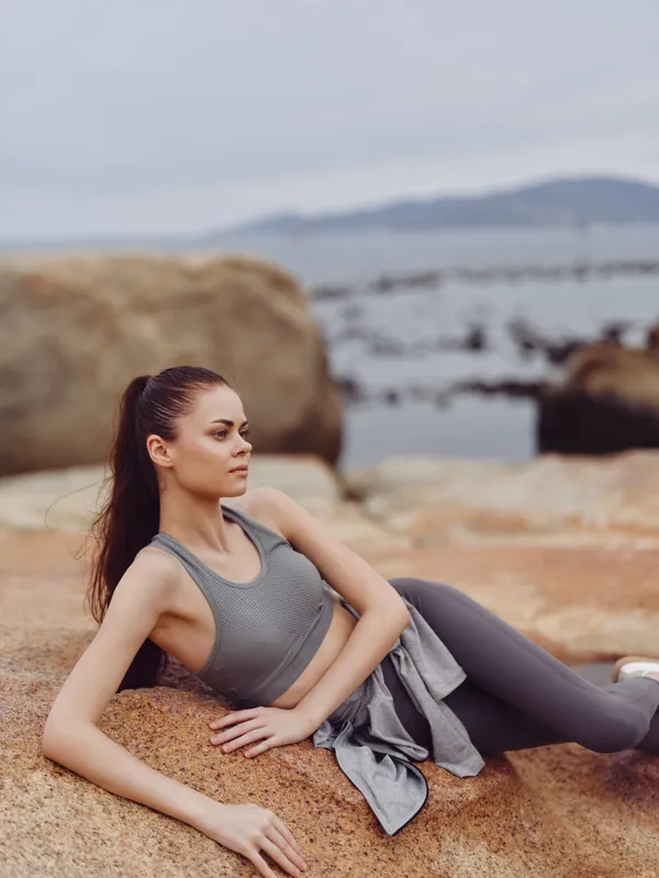 Young woman in athletic wear practicing yoga on rocky beach, showcasing athleticism and serenity in a nature setting with calm water in the background