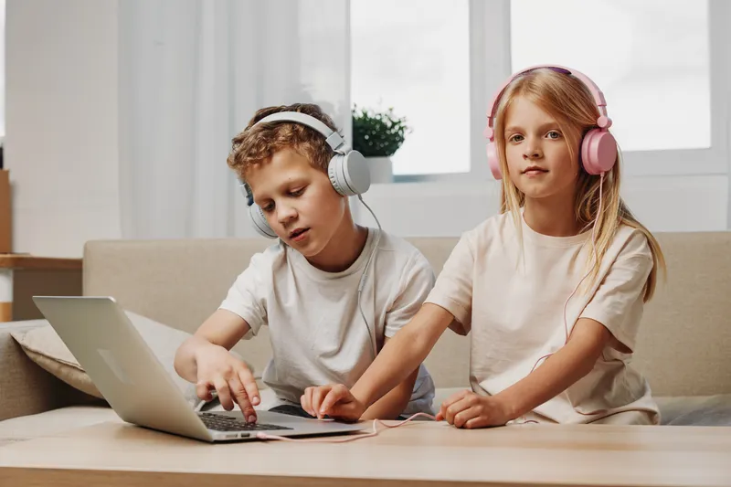 Two children wearing headphones use laptop at wooden table in bright room. The boy interacts with device while the girl watches. Concept of technology, education, and childhood.