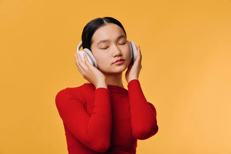 Young woman with closed eyes listening to music on headphones in red sweater against yellow background. Relaxed, serene, and focused on audio experience in minimal studio setting.