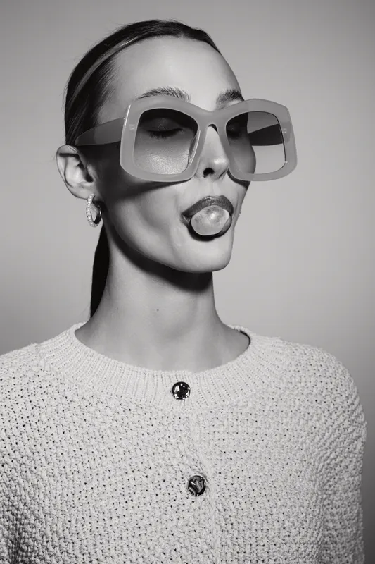 Stylish white woman with pale skin wears large square sunglasses and a textured sweater while blowing a bubblegum bubble in black and white studio portrait.