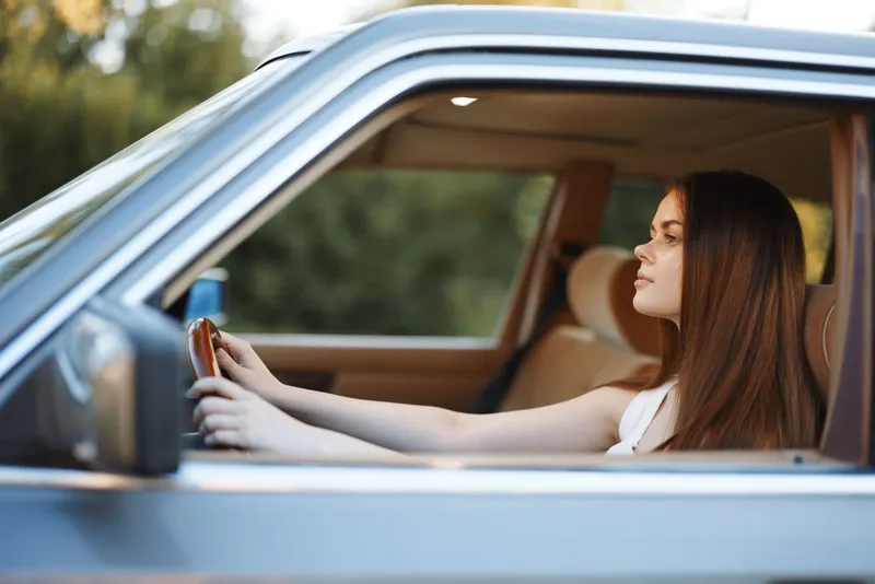 Young woman with long hair driving a vintage car outdoors, showcasing confidence and relaxation in a serene environment.