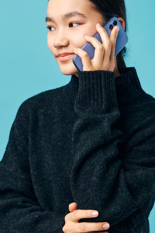 Young woman with dark hair wearing black sweater talking on smartphone against blue background. Female person closeup, casual style, communication and technology concept with smile and attention.
