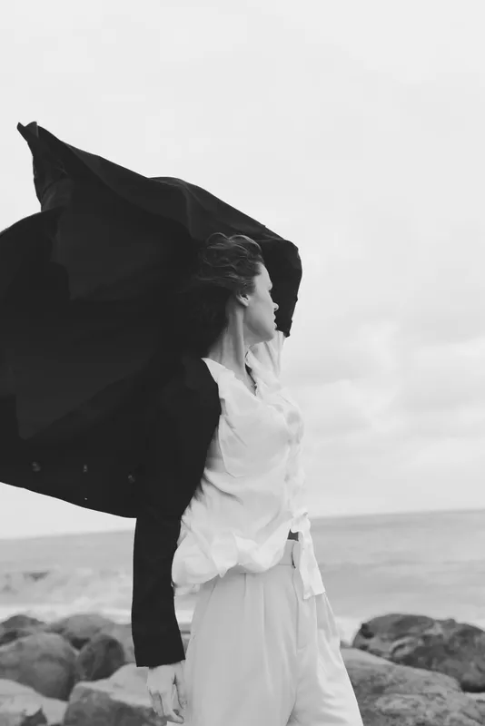 Wind sweeps a flowing cloth around a woman by the ocean, capturing movement, freedom, and a moody horizon in a monochrome frame, creating a dramatic seaside mood and graceful posture