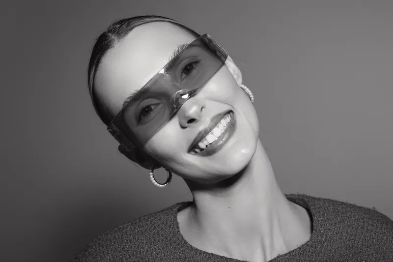 Smiling latina woman with medium skin tone wearing large sunglasses and hoop earrings. Portrait in black and white with soft lighting and casual clothing. Concept of style and happiness.