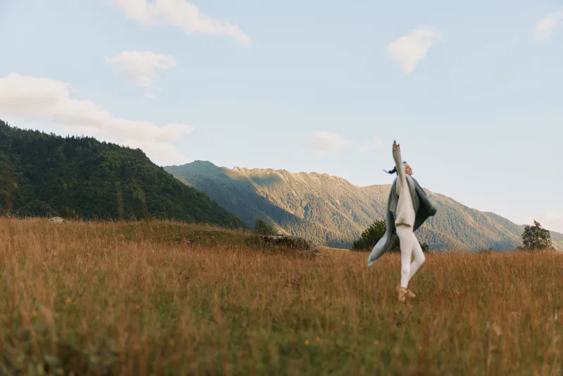 Woman in a wide field under a blue sky dances with a scarf, enjoying outdoor lifestyle, mountains in the distance, relaxed joyful mood, natural light and fresh air fills her moment.