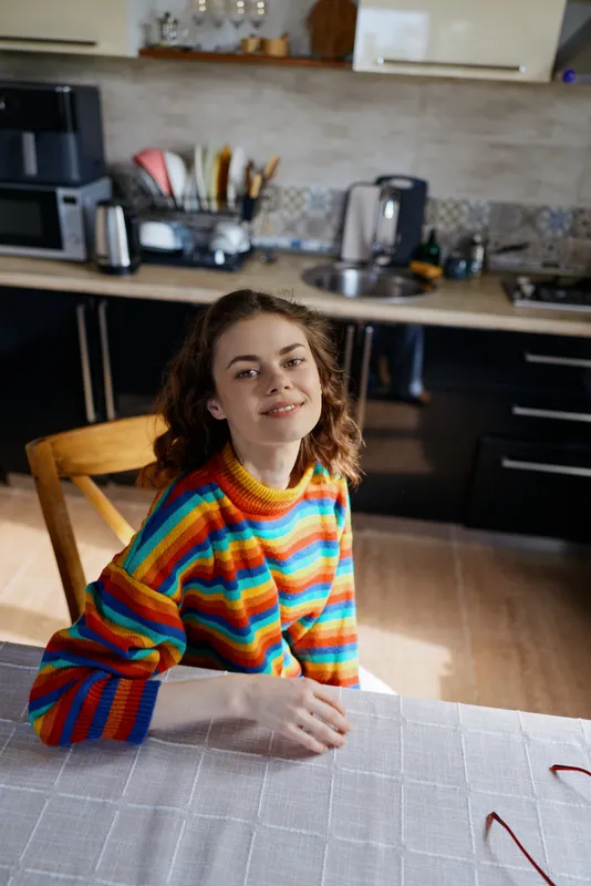Cheerful girl sits at a table in a bright kitchen, wearing a rainbow striped sweater, relaxed smile, natural light, domestic lifestyle moment, casual home portrait