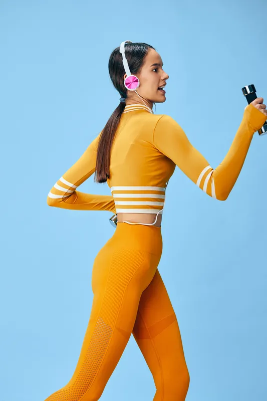 Woman fitness and sport theme, vibrant orange athletic wear, headphones, and bottle in hand mid workout against a blue studio backdrop, showcasing energy and focus.