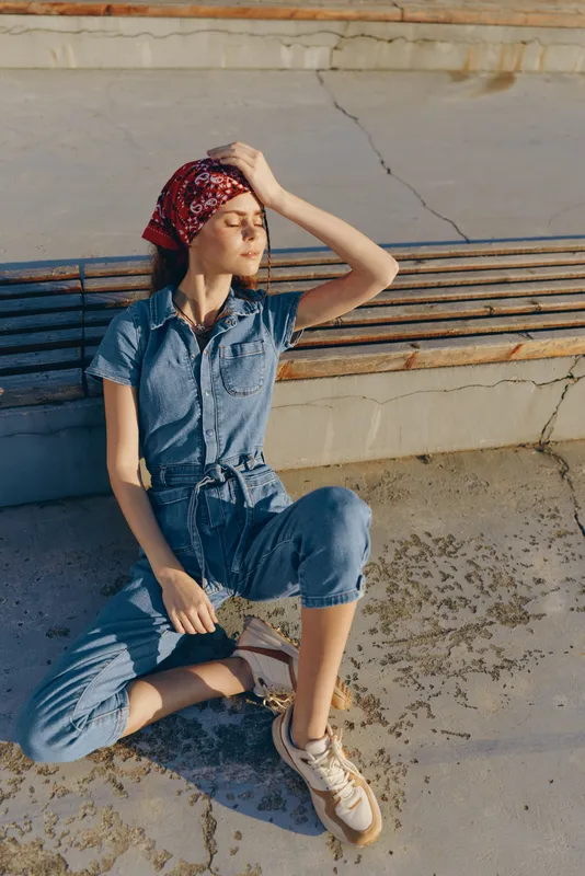 Lifestyle woman in modern boho-western denim street style relaxing outdoors with a red bandana and casual sneakers, captured in warm film color tones.
