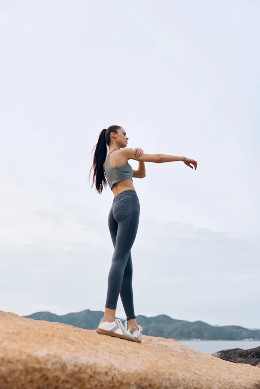 Fit young woman stretching outdoors in athletic wear by the sea at sunrise showcasing her healthy lifestyle, fitness, and motivation