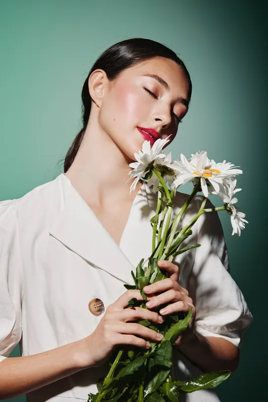 Beautiful young woman with dewy skin and glossy red lips holds white daisies while wearing a elegant white blouse. Soft studio lighting and pastel green background. Beauty fashion creative.