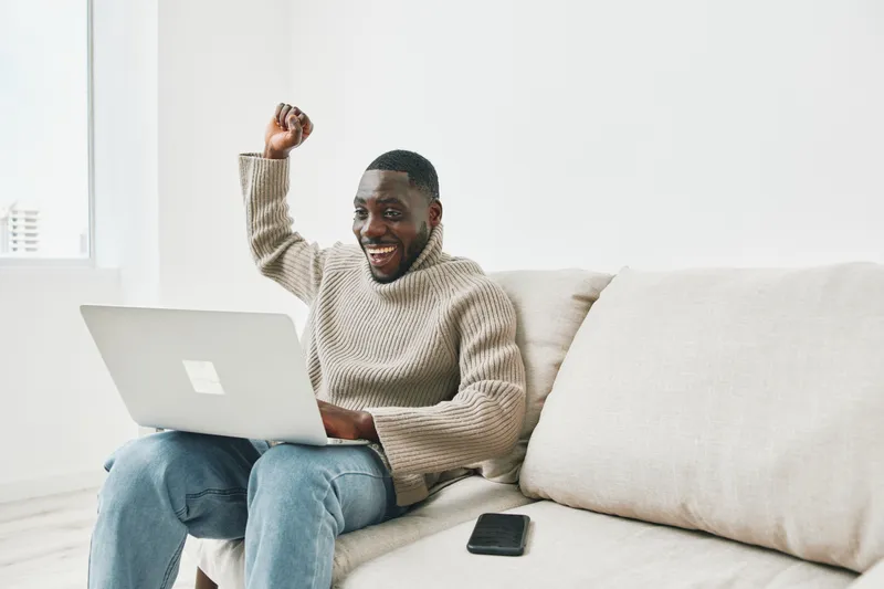 Happy Black man celebrating success while working on a laptop at home, wearing a cozy sweater, emphasizing positivity and productivity in a bright living space