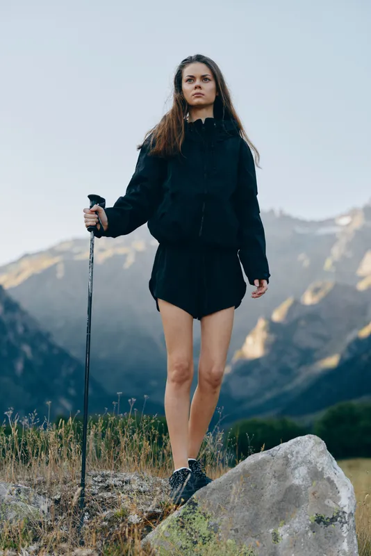 A lone hiker stands on a rocky outcrop in a mountainous landscape. She wears a dark jacket and shorts, gripping a trekking pole while looking ahead with focused determination.