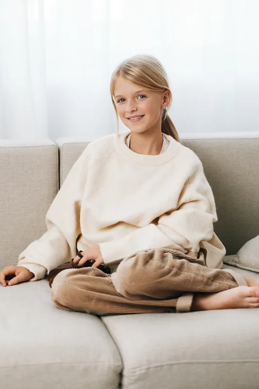 Young girl sits cross legged on a light sofa, wearing a cream sweater and brown pants, smiling softly. The image conveys cozy home comfort and a relaxed, candid moment.